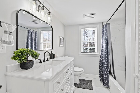 Une salle de bain moderne avec un meuble-lavabo blanc, un grand miroir, une plante en pot, des toilettes et une douche dotée d’un rideau à motifs.