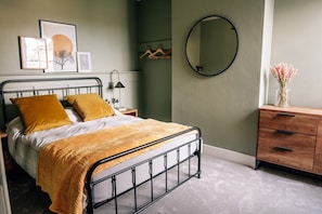 A bedroom with a metal bed frame, a wooden dresser, a mirror, and wall-mounted artwork. - Tranquility Parkside luxury 2 bed garden apartment (Worthing)