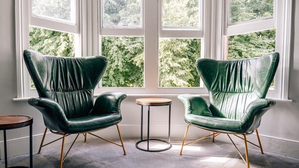 Dos sillones de cuero verde con patas de madera y una mesita redonda entre ellos, ubicados frente a una ventana con vista a árboles.
