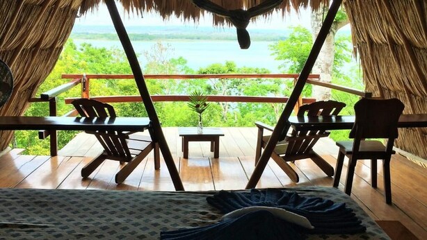 A room with a bed, a small table, and two chairs. The room has a view of a body of water and greenery. - Posada del Cerro (Flores)