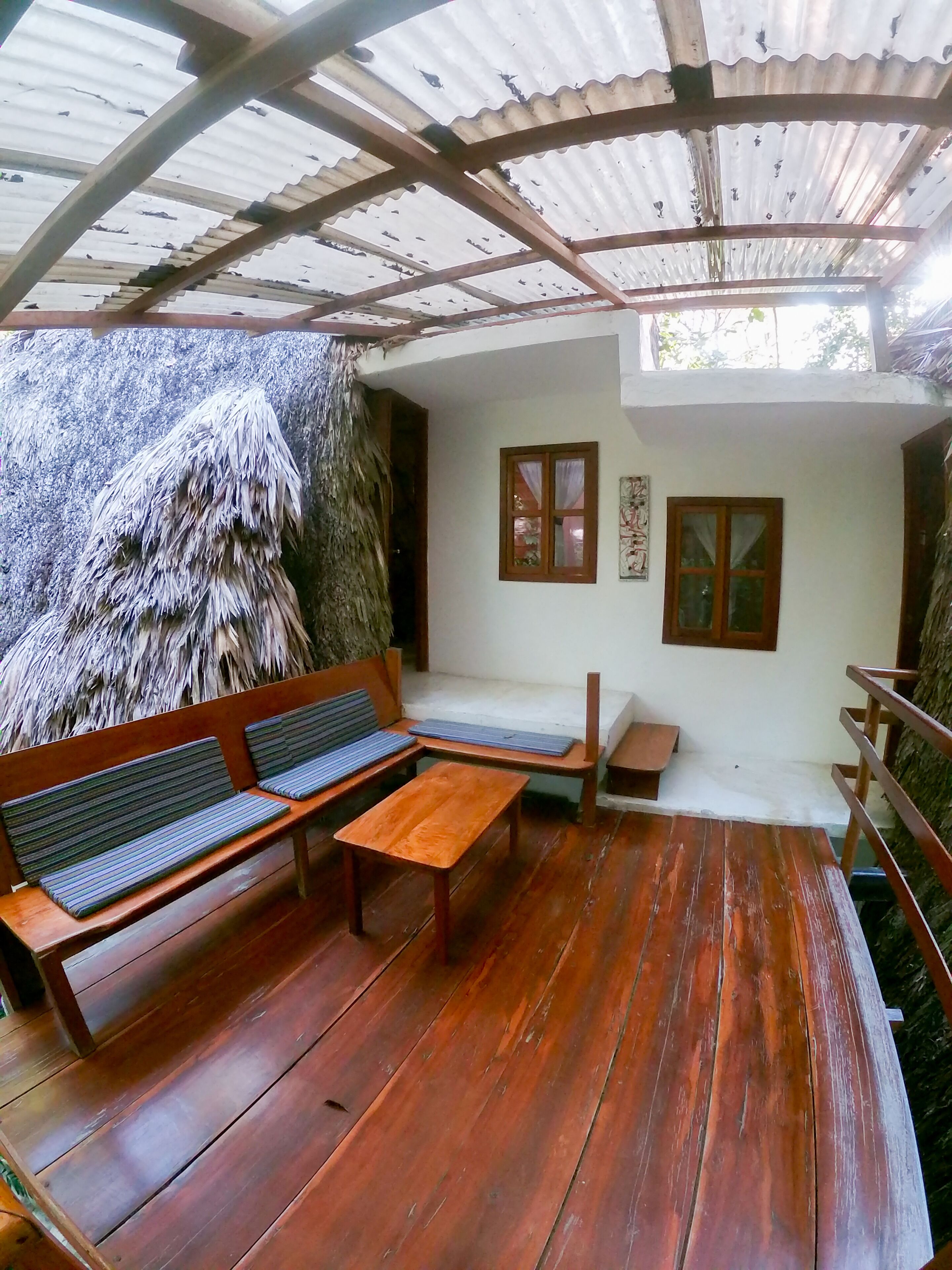 A wooden deck with a bench and a table, a covered area with a corrugated metal roof, and a wall with two windows.