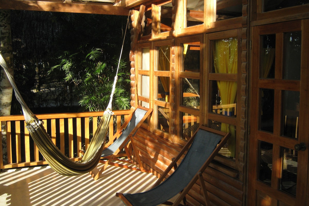 A wooden deck with a hammock and two lounge chairs, overlooking a tropical forest.