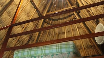 A thatched roof room with a wooden ladder, a mattress, and a ceiling fan.