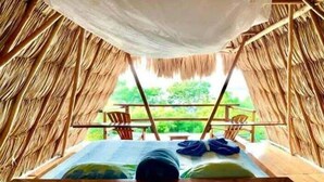 A thatched-roof tent with a bed and a table, a balcony with chairs, and a view of greenery outside. - Posada del Cerro (Flores)