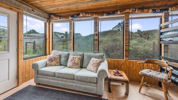 Ein gemütliches Zimmer mit Holzbalkendecke, einem grauen Sofa mit floralen Kissen, einem Holzstuhl und großen Fenstern mit Blick auf einen Hügel und ein Gebäude.
