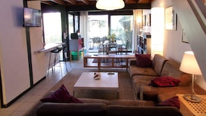 A cozy living room with a sofa, coffee table, and a TV mounted on the wall. There is a dining area with a table and chairs, and a staircase leading to another level. - Beautiful 67 m² duplex with garden.golf view (Moliets-et-Maa)