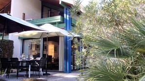 An outdoor dining area with tables and chairs, a large umbrella, and a building with blue and white exterior walls. - Beautiful 67 m² duplex with garden.golf view (Moliets-et-Maa)