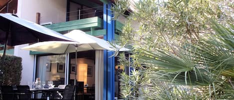 Un espace extérieur aménagé pour les repas, avec des tables et des chaises, un grand parasol, et un bâtiment dont les murs extérieurs sont peints en bleu et en blanc.