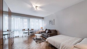 A modern, well-lit room with a wooden floor, a sofa, a bed, a dining table with chairs, and a large window with sheer curtains. - Athénée 36 by Homenhancement  (Geneva)