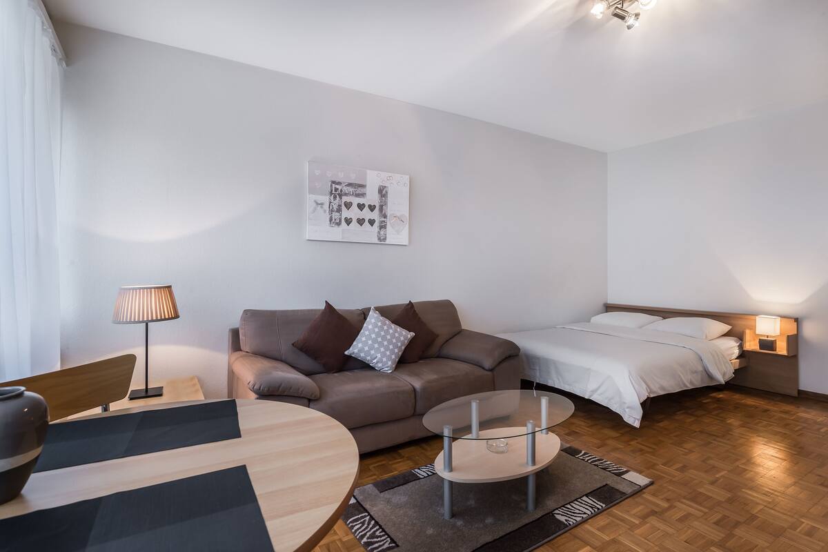A modern, well-lit hotel room with a sofa, a round coffee table, a bed, and a wall-mounted lamp.