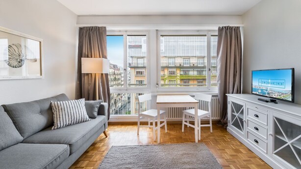 A modern living room with a grey sofa, white dining table, and chairs, a TV on a cabinet, and large windows with curtains. - Athénée 36 by Homenhancement  (Geneva)