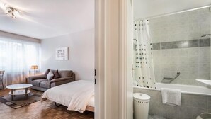 A bedroom with a bed, a sofa, a coffee table, and a window with curtains. - Athénée 36 by Homenhancement  (Geneva)