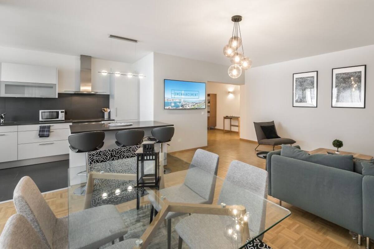 A modern living room with a glass dining table, grey sofas, and a kitchen in the background.