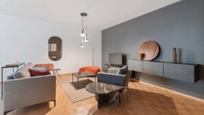 A modern living room with a grey sofa, a blue armchair, a round coffee table, and a TV on a stand. - Athénée 36 by Homenhancement  (Geneva)