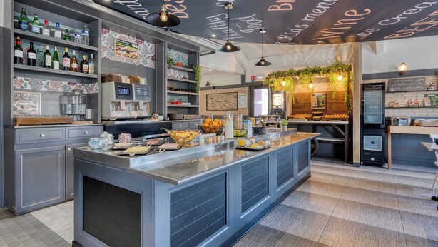 A modern bar area with a central counter, shelves with bottles, and a menu wall. - B&B Hotel Bordeaux Mérignac Cadera Aéroport (Merignac)