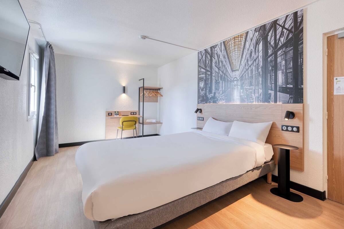A modern hotel room with a large bed, a desk, a chair, a lamp, and a wall-mounted artwork.