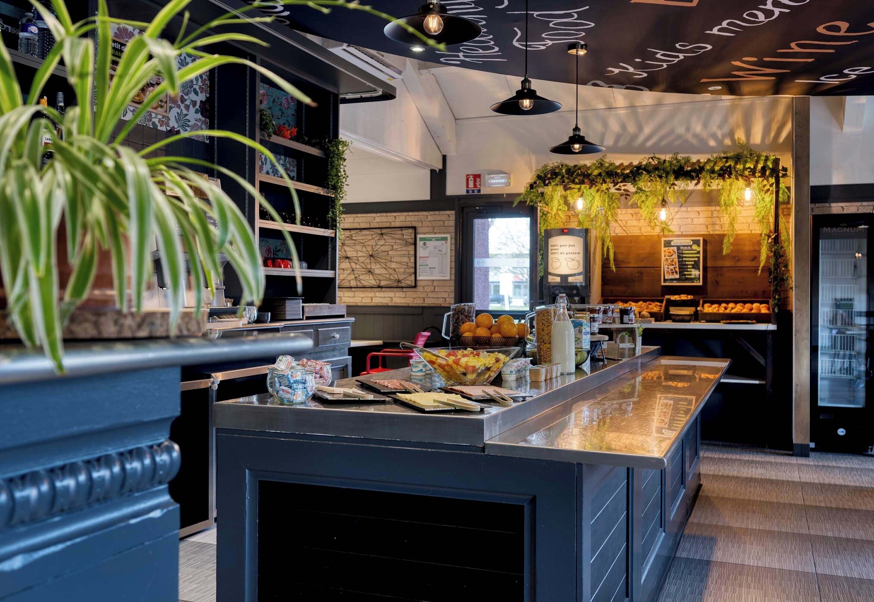 A modern bar area with a central counter, shelves with various items, hanging plants, and a menu board.