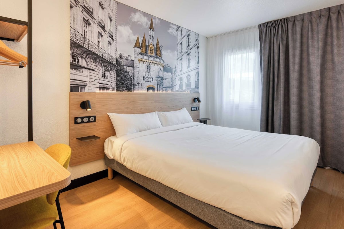 A hotel room with a large bed, a wooden desk, a small table, a headboard with controls, and a mural of a castle.