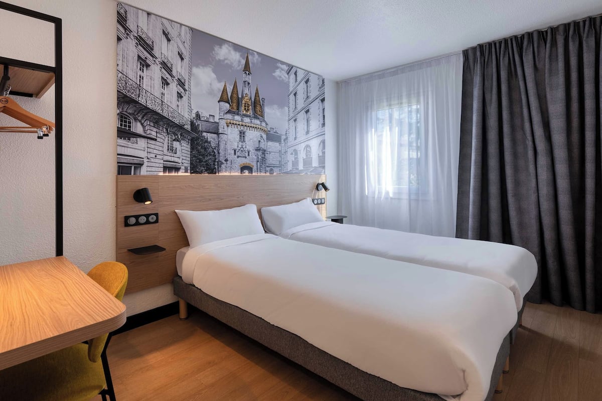 A hotel room with two beds, a desk, a chair, a wall-mounted shelf, and a large window with curtains.