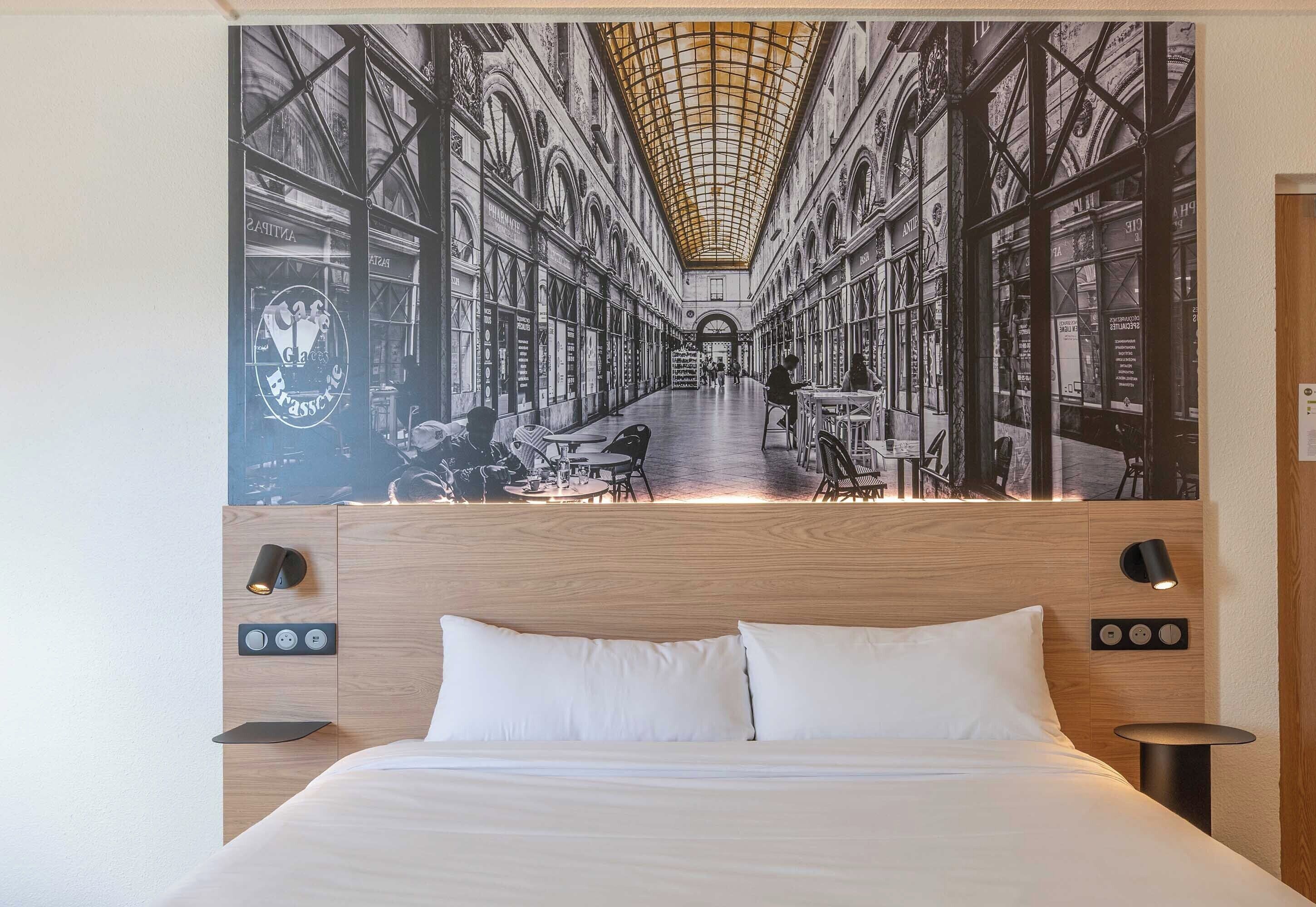A hotel room with a large mural of a train station, a wooden headboard, a bed with white bedding, and bedside tables with lamps.