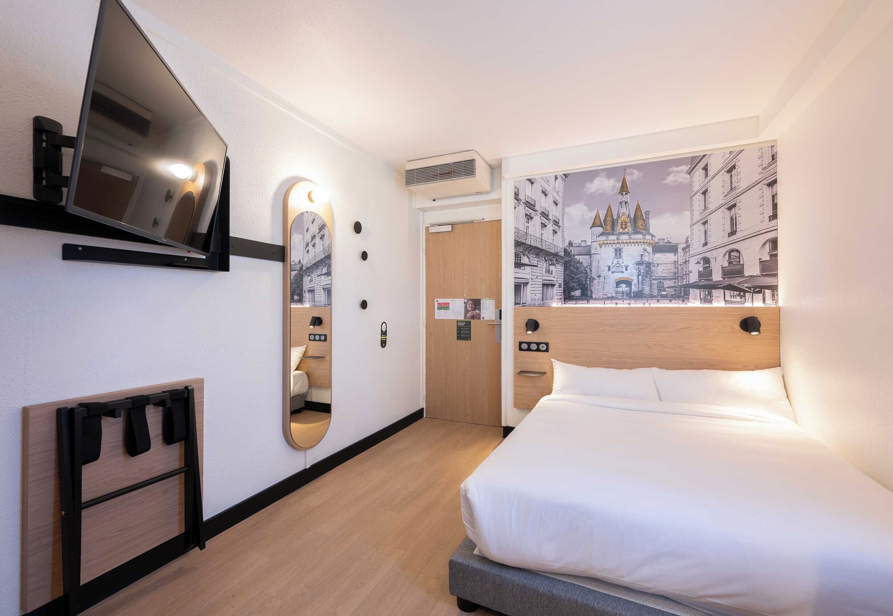 A hotel room with a bed, a mounted TV, a mirror, a wooden headboard, and a wall-mounted air conditioner.
