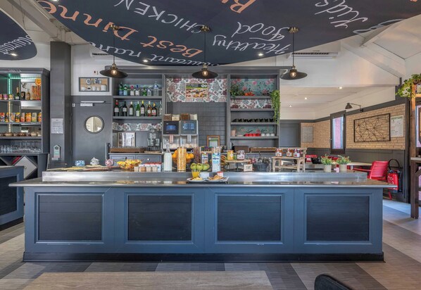 Una cafetería moderna con una barra central tipo isla, estanterías con bebidas y aperitivos, y un techo decorativo con texto.