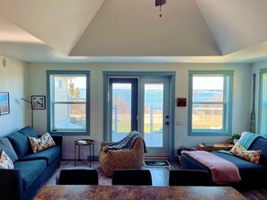 A living room with a sofa, armchair, and a table. There are three windows with blinds, a ceiling fan, and a view of the outdoors.