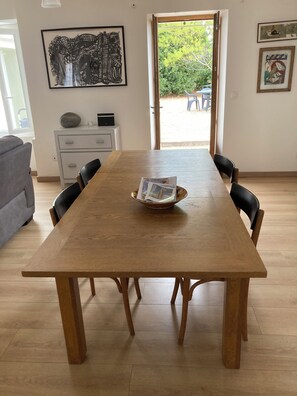 Ein Essbereich mit Holztisch, Stühlen und einer Schale mit Zeitschriften. An den Wänden befinden sich gerahmte Kunstwerke und ein Blick ins Freie durch eine Tür.