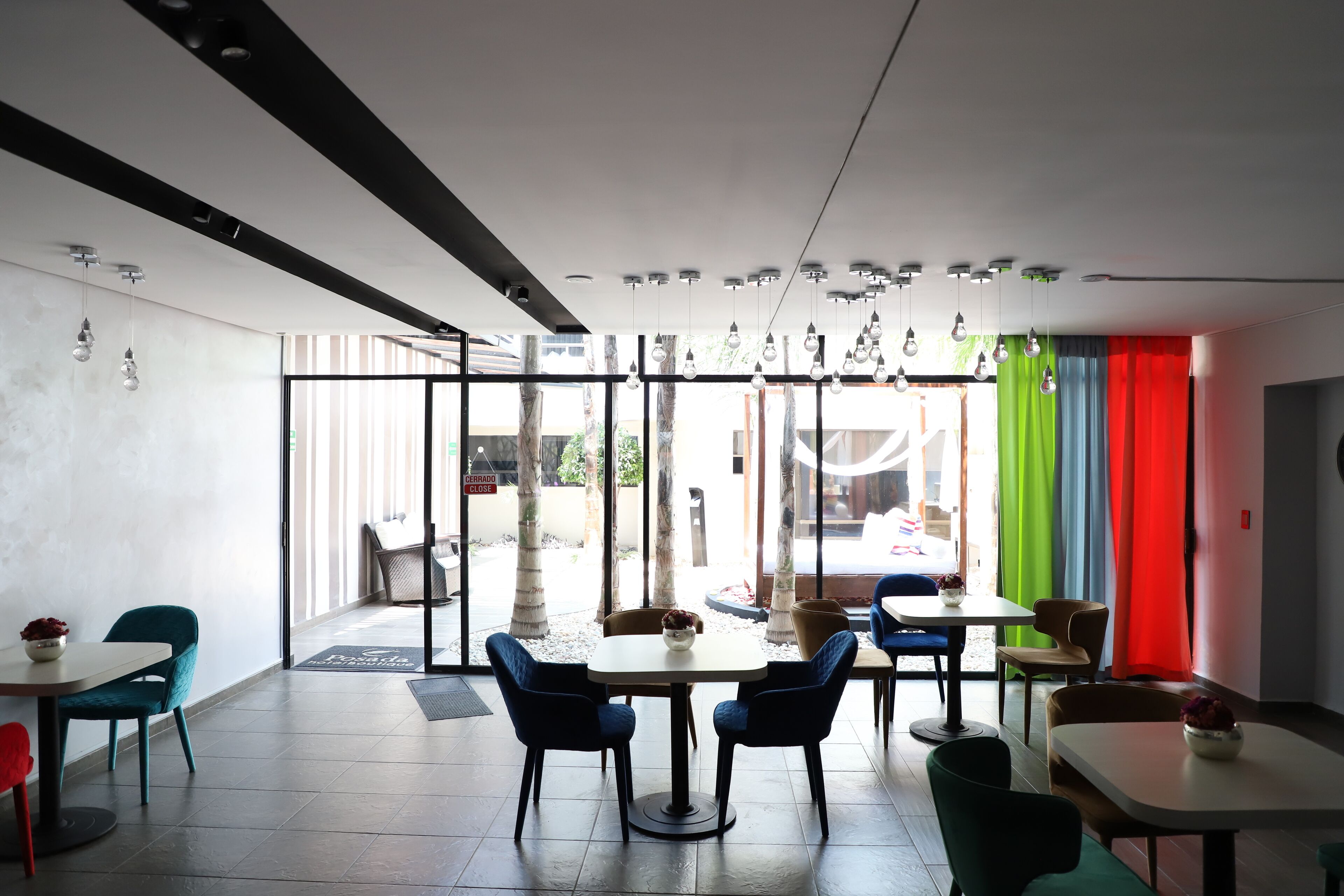 A modern interior with a dining area featuring round tables and blue chairs, large windows with colorful curtains, and hanging light fixtures.