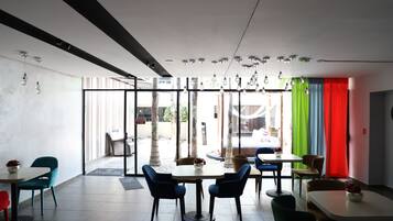 A modern interior with a dining area featuring round tables and blue chairs, large windows with colorful curtains, and hanging light fixtures.