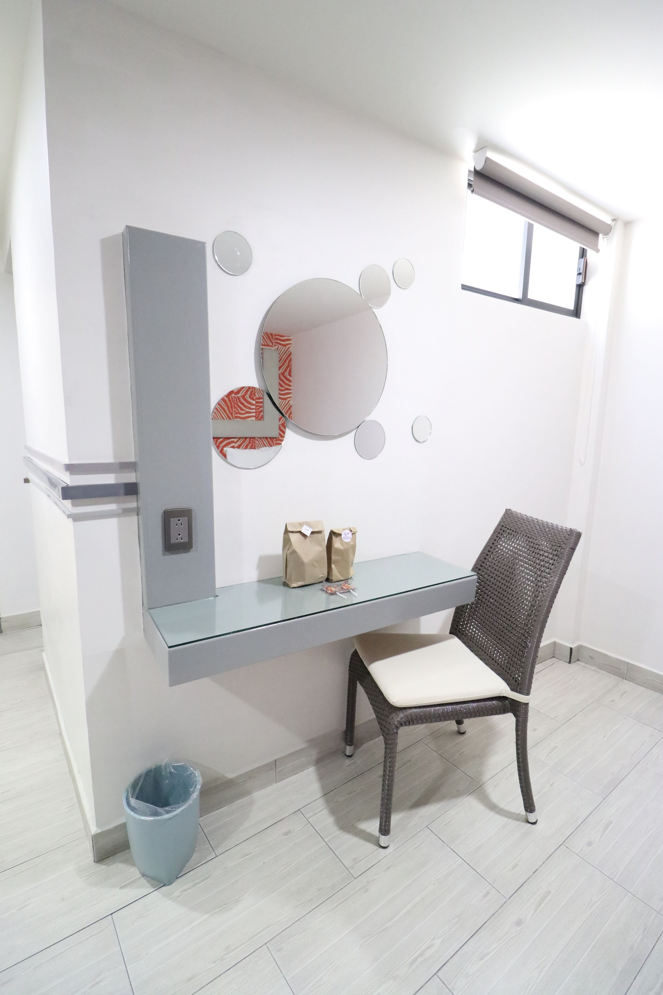 A room with a glass desk, a wicker chair, a wall-mounted mirror, and a small trash bin.