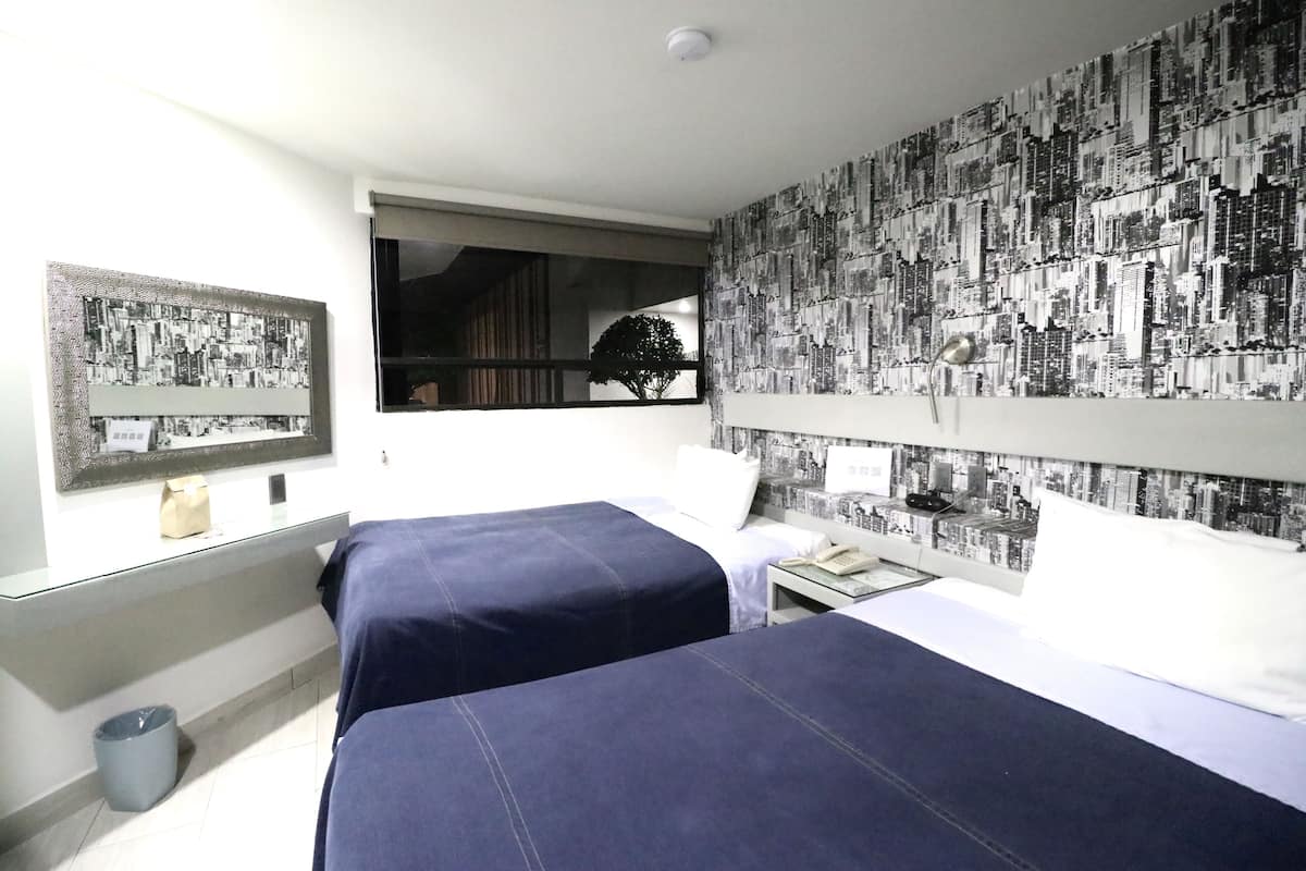 Habitación de hotel con dos camas, papel tapiz con vista a la ciudad, un cuadro enmarcado, una ventana que da a un árbol y una mesita con un teléfono.