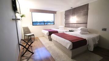 A modern bedroom with two beds, a large window, a chair, and a flat-screen TV mounted on the wall.