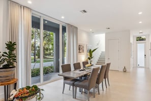 A modern dining area with a table and chairs, large windows with curtains, and a staircase leading to another level.