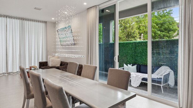 Een moderne eetruimte met een grote tafel en stoelen, een bank, en uitzicht op een groene haag en bomen door grote glazen schuifdeuren.