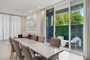 A modern dining area with a large table and chairs, a sofa, and a view of a green hedge and trees through large sliding glass doors.