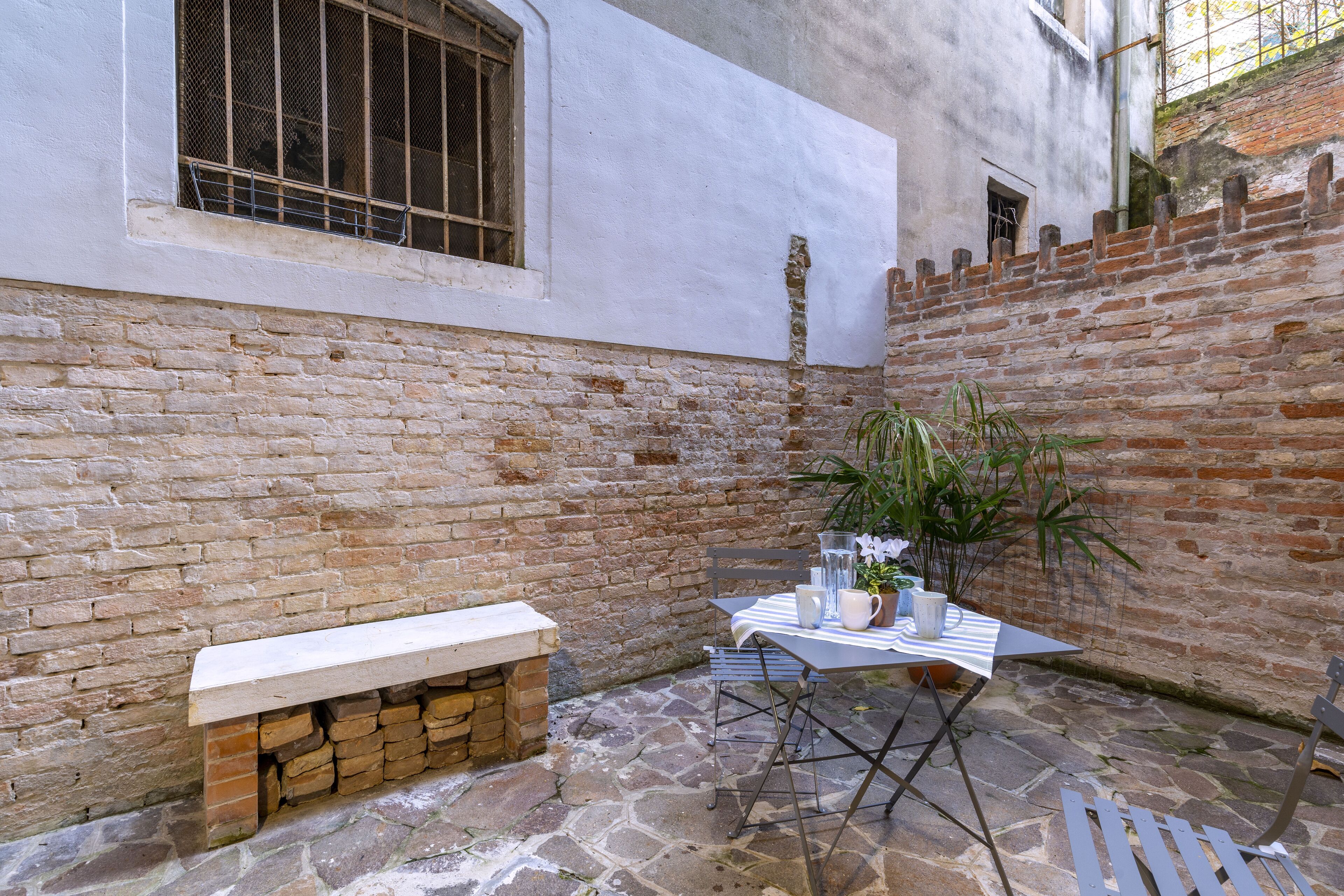 Un patio con suelo de piedra, una banca, una mesa con mantel a rayas y una planta en maceta.
