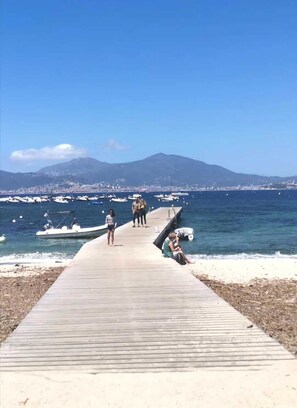 A wooden pier leading to a beach with boats in the water and a mountain in the background.