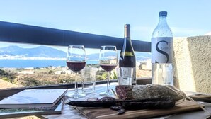 A table with two glasses of red wine, a bottle of water, and a loaf of bread on a balcony overlooking a scenic view of the sea and mountains.