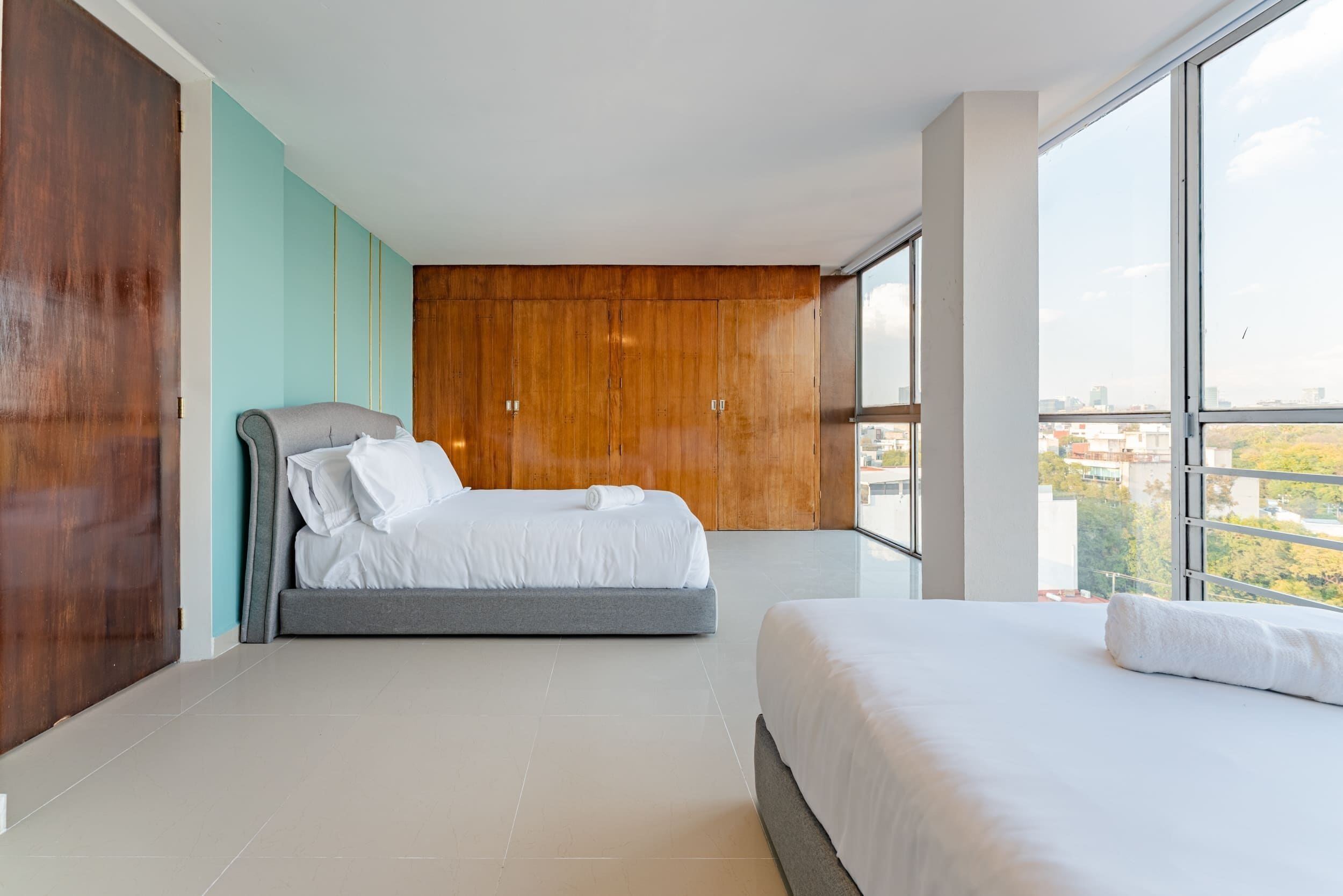 A modern bedroom with a large bed, a smaller bed, and a view of the city through large windows.