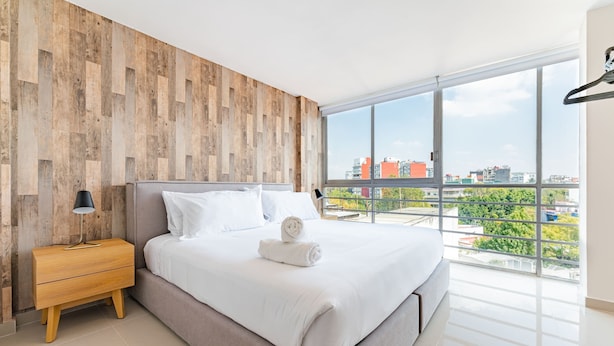 Une chambre moderne avec un grand lit, un lambris en bois, une table de chevet, une lampe et une vue sur la ville grâce à de grandes fenêtres.