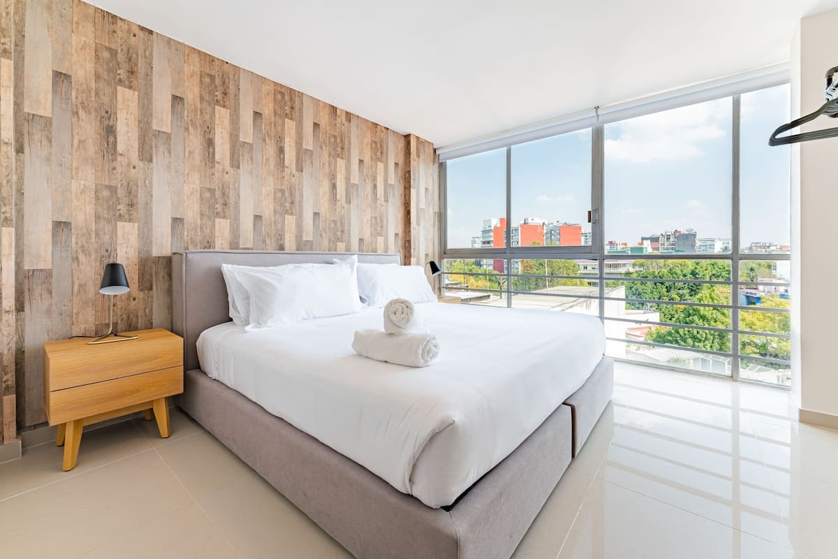 A modern bedroom with a large bed, wooden wall paneling, a nightstand, a lamp, and a view of the city through large windows.
