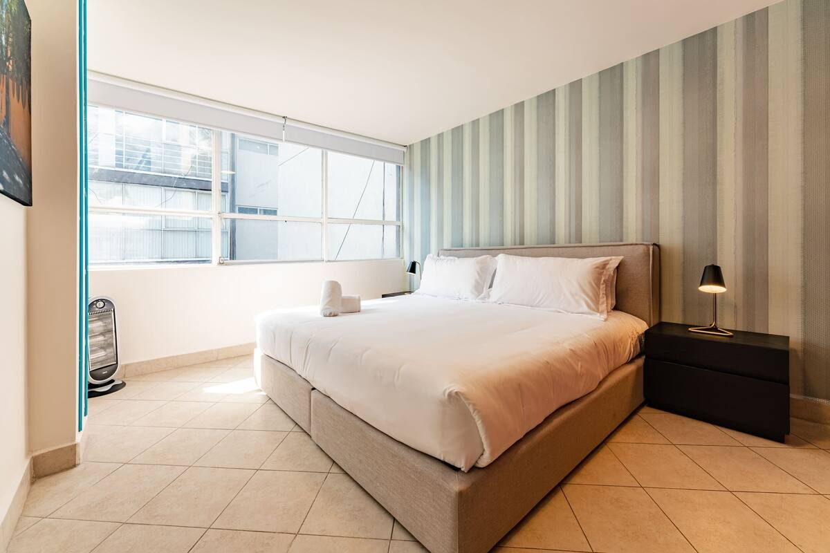 A bedroom with a large bed, a bedside table with a lamp, a window, and a heater.