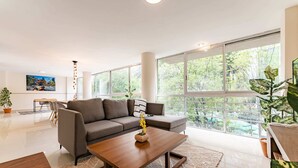 Un salon moderne avec un canapé d’angle, une table basse en bois et de grandes fenêtres donnant sur un paysage verdoyant.