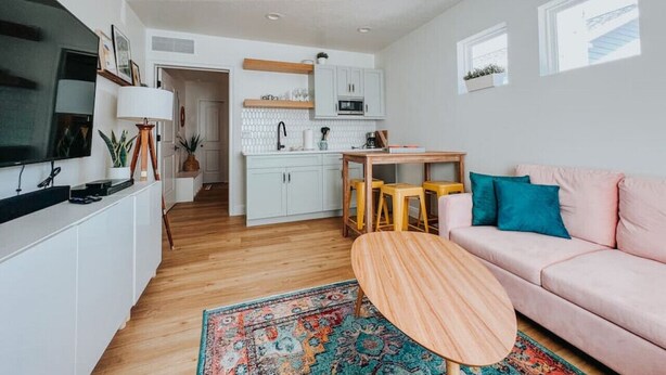 Una sala moderna con un sofá rosa, una mesa de centro de madera y un televisor sobre un mueble blanco. Se observa una zona de cocina con armarios blancos y un microondas.