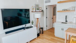 Una cocina moderna con armarios blancos, piso de madera, un televisor en la pared y un pequeño comedor con una mesa y sillas.