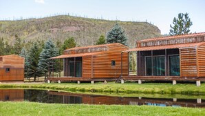 A row of wooden cabins with large windows, set against a backdrop of a hill and a body of water. - Riverside Suite Casita 1 (Greer)