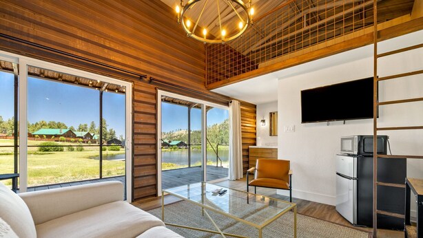 A modern living room with a glass coffee table, a sofa, a TV, and a kitchen area with a refrigerator and microwave. - Riverside Suite Casita 6 (Greer)