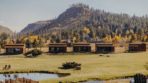Un hilera de cabañas rústicas con ventanas amplias, ubicadas en un campo de césped con montañas al fondo.