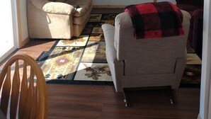 Una sala de estar con un sofá beige, un sillón gris con una manta roja y negra, un ventilador de techo, un televisor de pantalla plana y una puerta con marco de madera.
